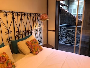 Un dormitorio con cama, almohadas, cabecera de metal decorativa y vista a un balcón con barandilla.