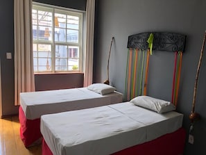Dos camas individuales con sábanas blancas y reposapiés rojos en una habitación con un ventanal grande y un cabecero decorativo de metal.