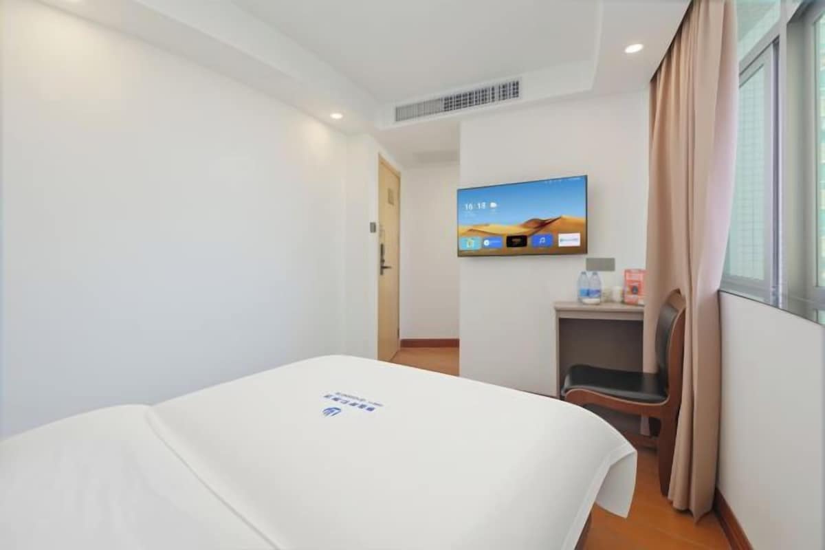 A hotel room with a bed, a television mounted on the wall, a small desk with a chair, and a window with curtains.