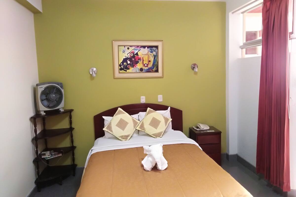 Habitación de hotel con una cama, una mesita de noche, un ventilador de pared, un cuadro y una ventana con cortinas rojas.