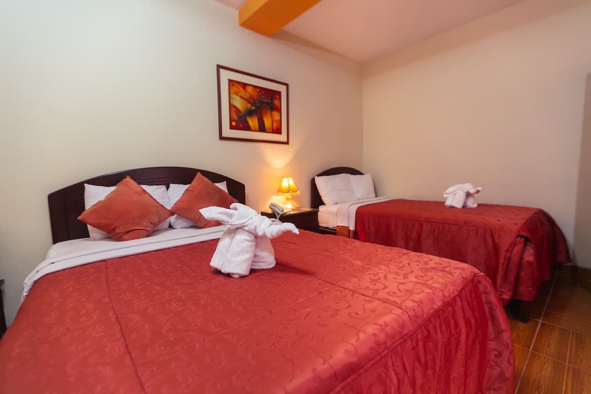 Habitación de hotel con dos camas, cada una con ropa de cama y almohadas rojas, una mesita de noche con lámpara y un cuadro en la pared.