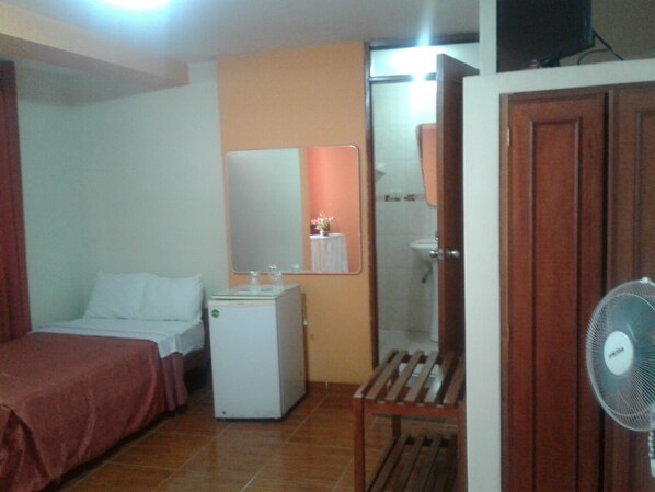 Quarto de hotel com cama, pequeno frigorífico, espelho, chuveiro e ventoinha.