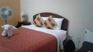 Habitación de hotel con cama, mesita de noche, lámpara, ventilador, un peluche y escritorio con silla.