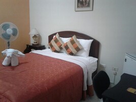 Quarto de hotel com cama, mesa de cabeceira, abajur, ventilador, um brinquedo de pelúcia e escrivaninha com cadeira.