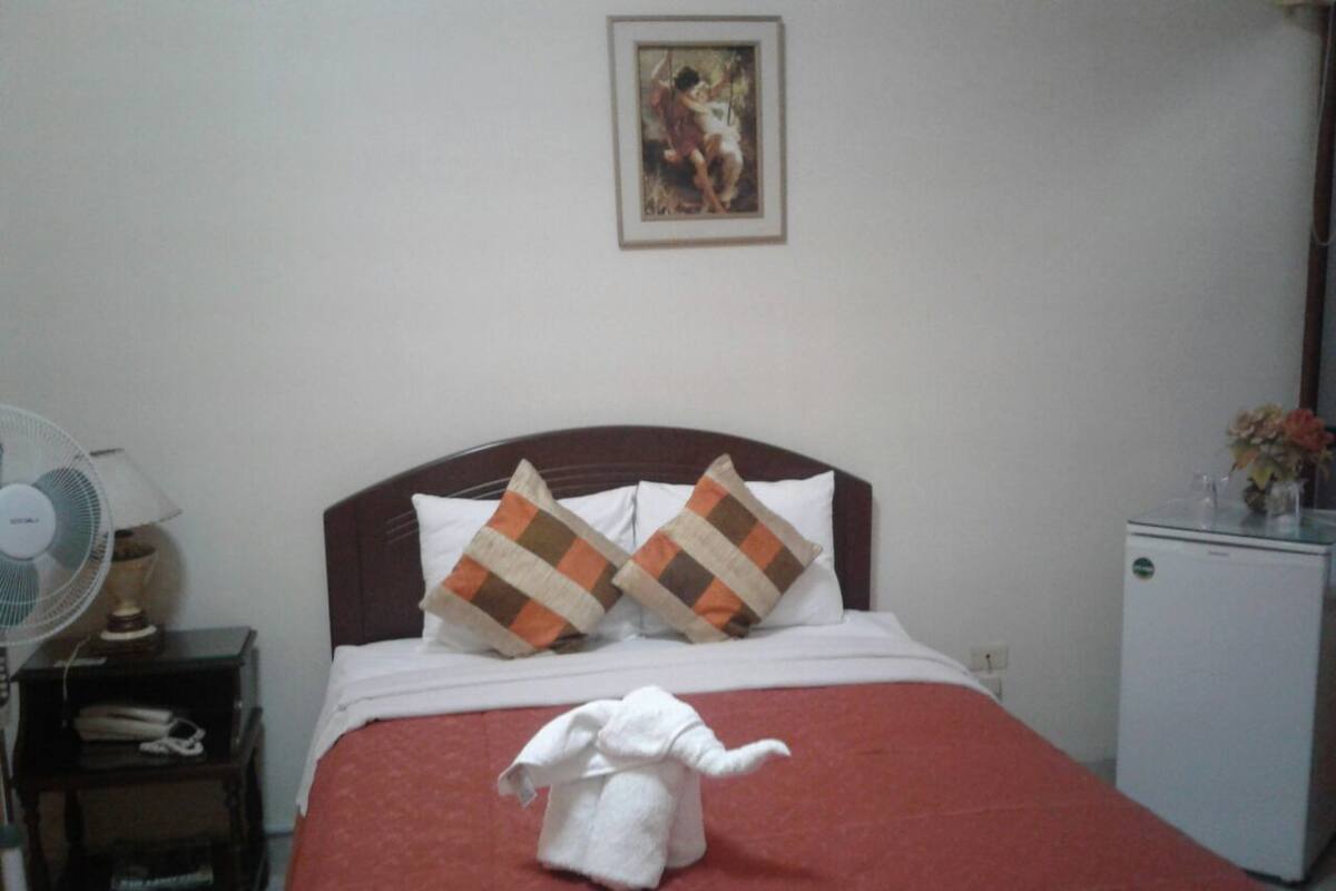 Habitación de hotel con cama, almohadas, ventilador, un refrigerador pequeño y una toalla con forma de elefante.