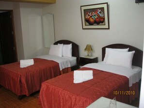 Quarto de hotel com duas camas, cada uma com colcha vermelha e almofadas brancas, uma mesa de cabeceira com abajur e um quadro na parede.
