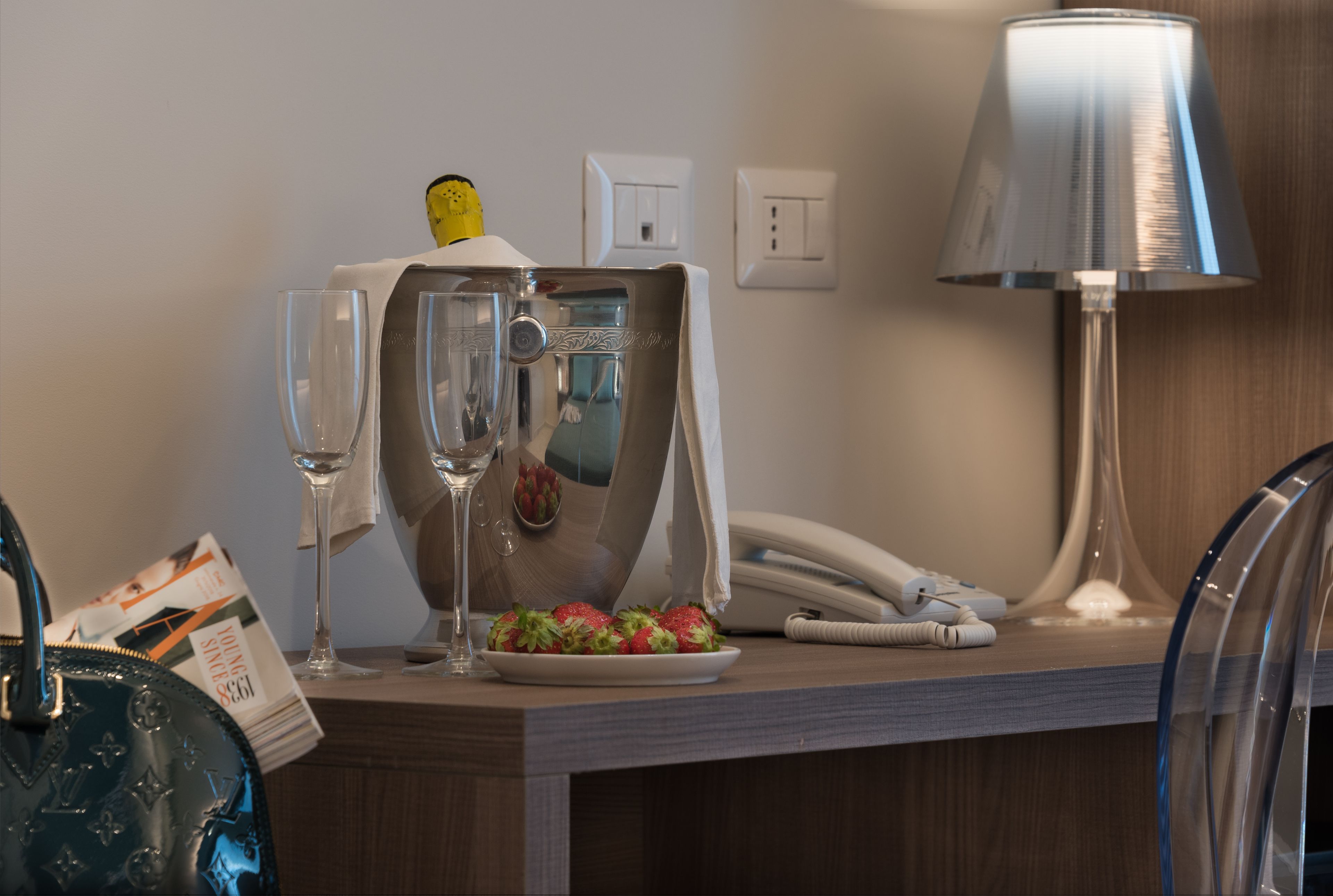 A hotel room counter with a minibar, a telephone, a lamp, and a plate of strawberries.