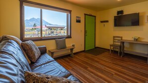 Una habitación con piso de madera, un sofá con almohadas, un banco, una puerta verde, un televisor montado en la pared y vista a las montañas a través de una ventana.