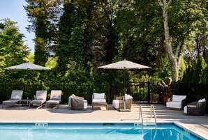 Zona junto a la piscina con sillones de descanso y sombrillas, rodeada por una valla y árboles.