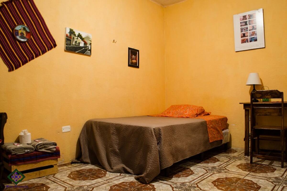 A small, cozy room with a bed, a desk, and a chair. There are pictures on the wall and a lamp on the desk.