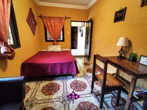 A room with a bed, a desk, and a chair. There is a window with curtains, a door, and a wall with pictures.