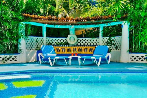 Un espace piscine agrémenté d’un ensemble de chaises longues bleues et jaunes placé sous une pergola, le tout entouré d’une végétation luxuriante.