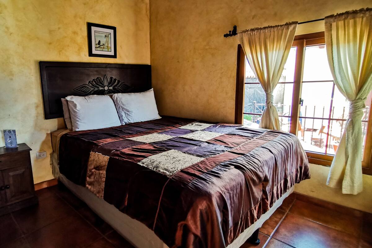 Un dormitorio con una cama, una mesita de noche, un cuadro enmarcado en la pared y una ventana con cortinas.
