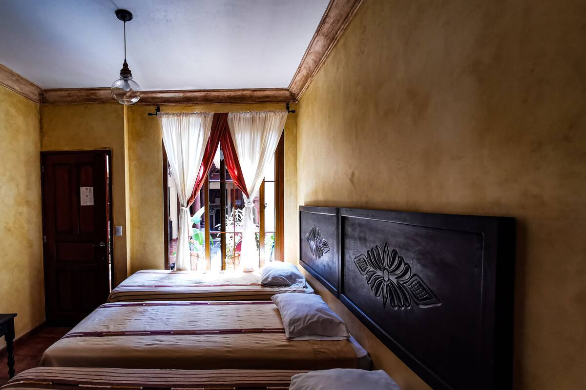 Habitación de hotel con dos camas, cabecera de madera y vista al exterior a través de una ventana con cortinas rojas.