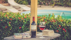 A bottle of wine, a glass of wine, a book, and a hat on a table by a poolside. - Pousada Vila do Mar (Sao Jose de Ribamar)