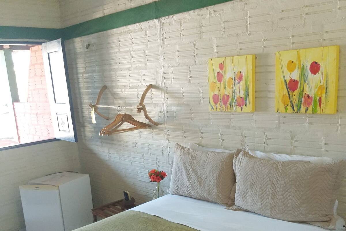 A hotel room with a bed, a green blanket, a white pillow, a wooden hanger, and two paintings on the wall.