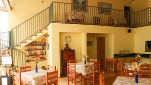 A spacious dining area with multiple wooden tables and chairs, a staircase leading to an upper level, and a bookshelf filled with books and decorative items. - Pousada Vila do Mar (Sao Jose de Ribamar)