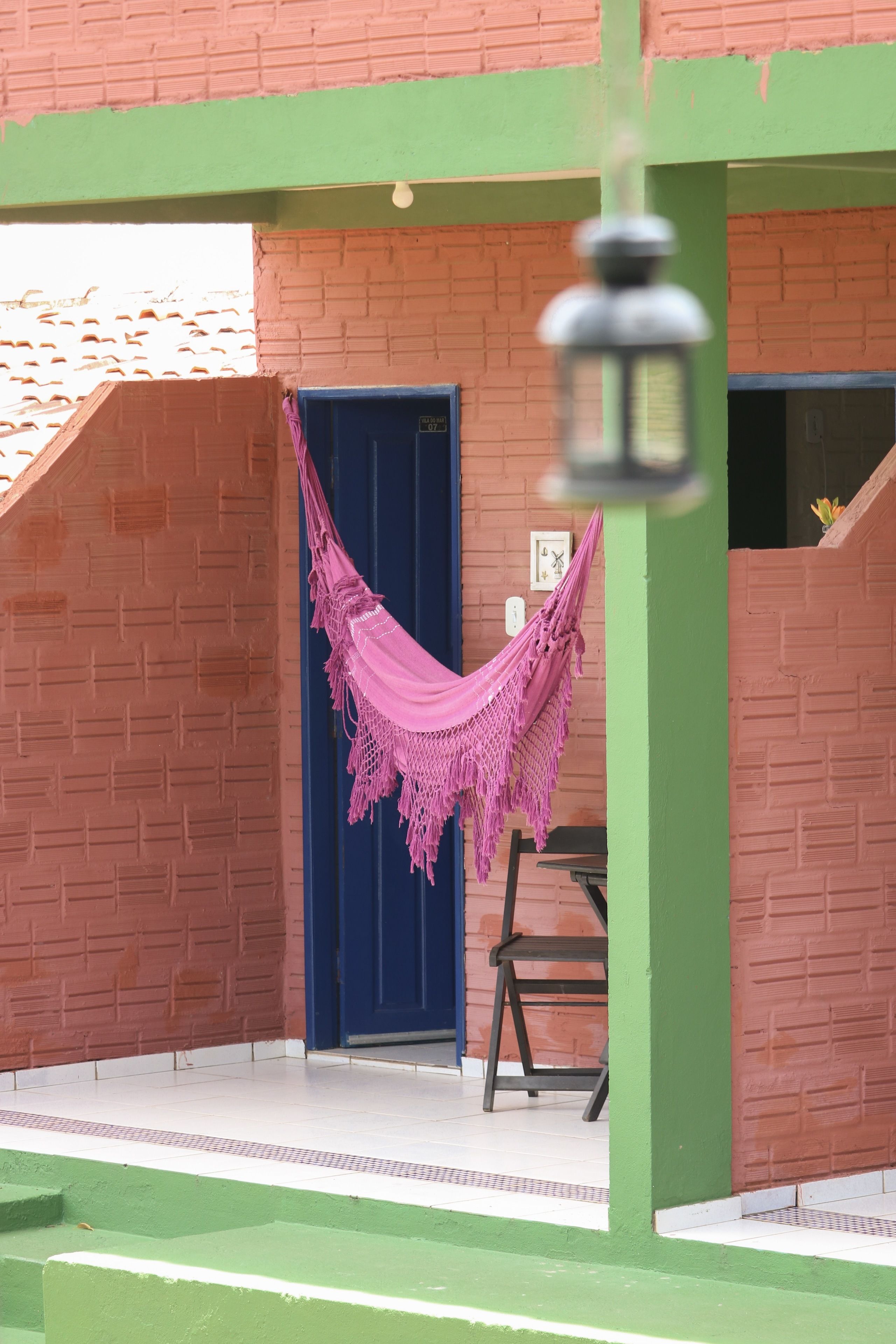 A brick building with a blue door, a hammock hanging from a green structure, and a chair outside.