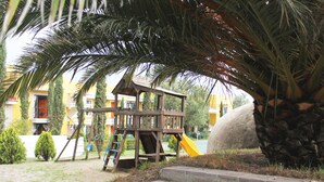 A wooden play structure with a slide, surrounded by palm trees and a building in the background. - Quinta Huizache (Nopaltepec)