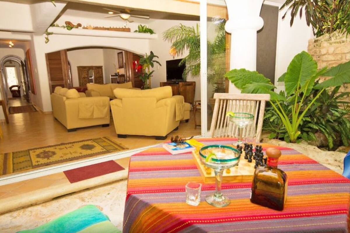 Ein Wohnzimmer mit einer Couch, einem Esstisch mit farbigem Tischläufer und einer Zimmerpflanze in der Ecke.