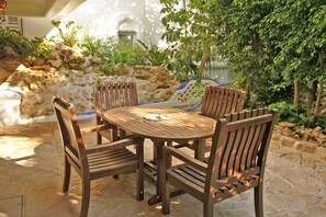 Una mesa de madera redonda con cuatro sillas en un espacio exterior iluminado por el sol, con suelo de piedra y una pared de piedra cubierta de vegetación.