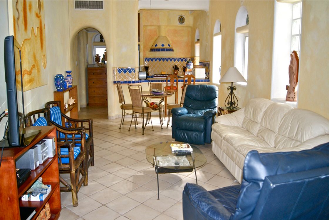 A living room with a sofa, chairs, a coffee table, and a TV. There is a kitchen area with a tiled backsplash and a dining table.