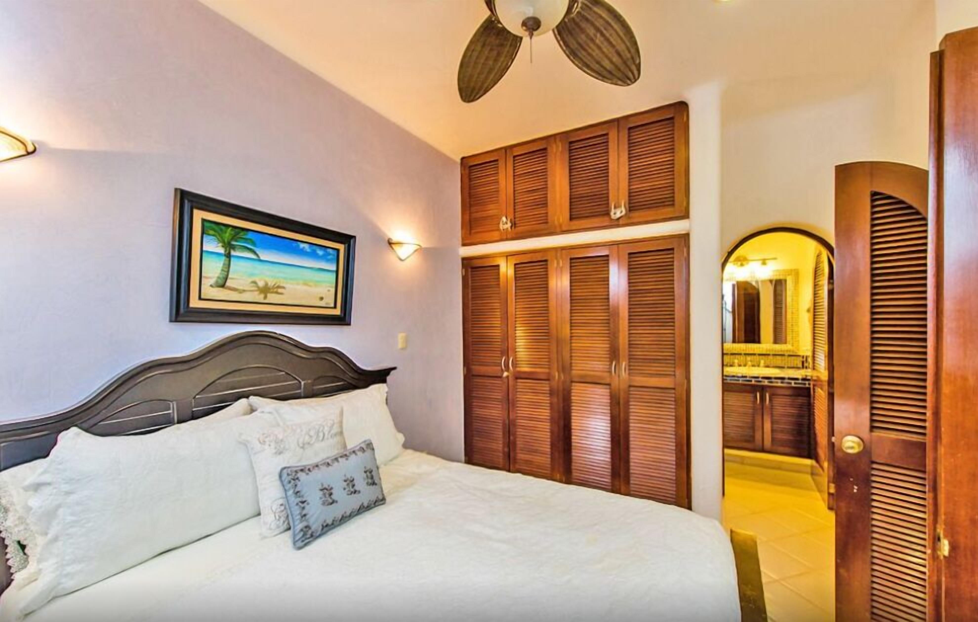 A bedroom with a bed, wooden wardrobe, a ceiling fan, a framed picture, and a mirror.
