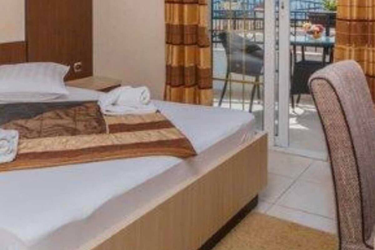 Quarto de hotel com uma cama grande, uma cadeira e vista para a cidade através de uma porta de vidro.