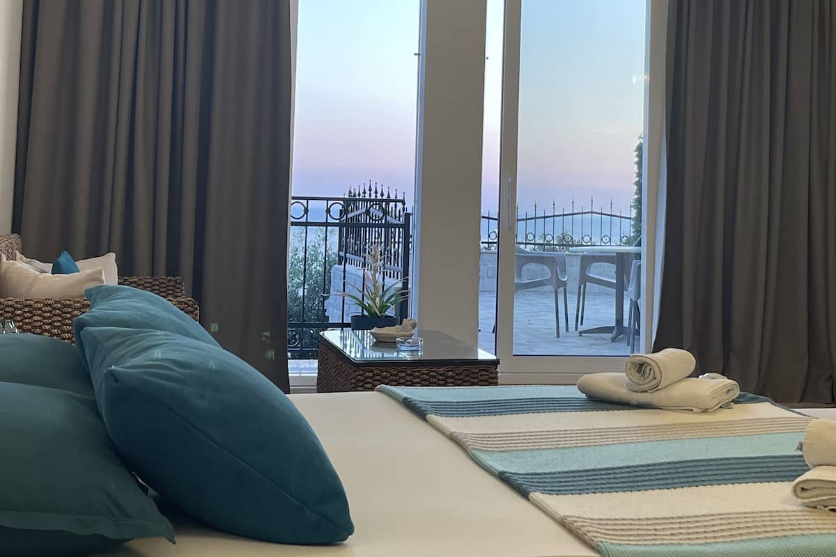 Quarto de hotel com uma cama grande, vista para o mar através de janelas do chão ao teto e um candelabro.