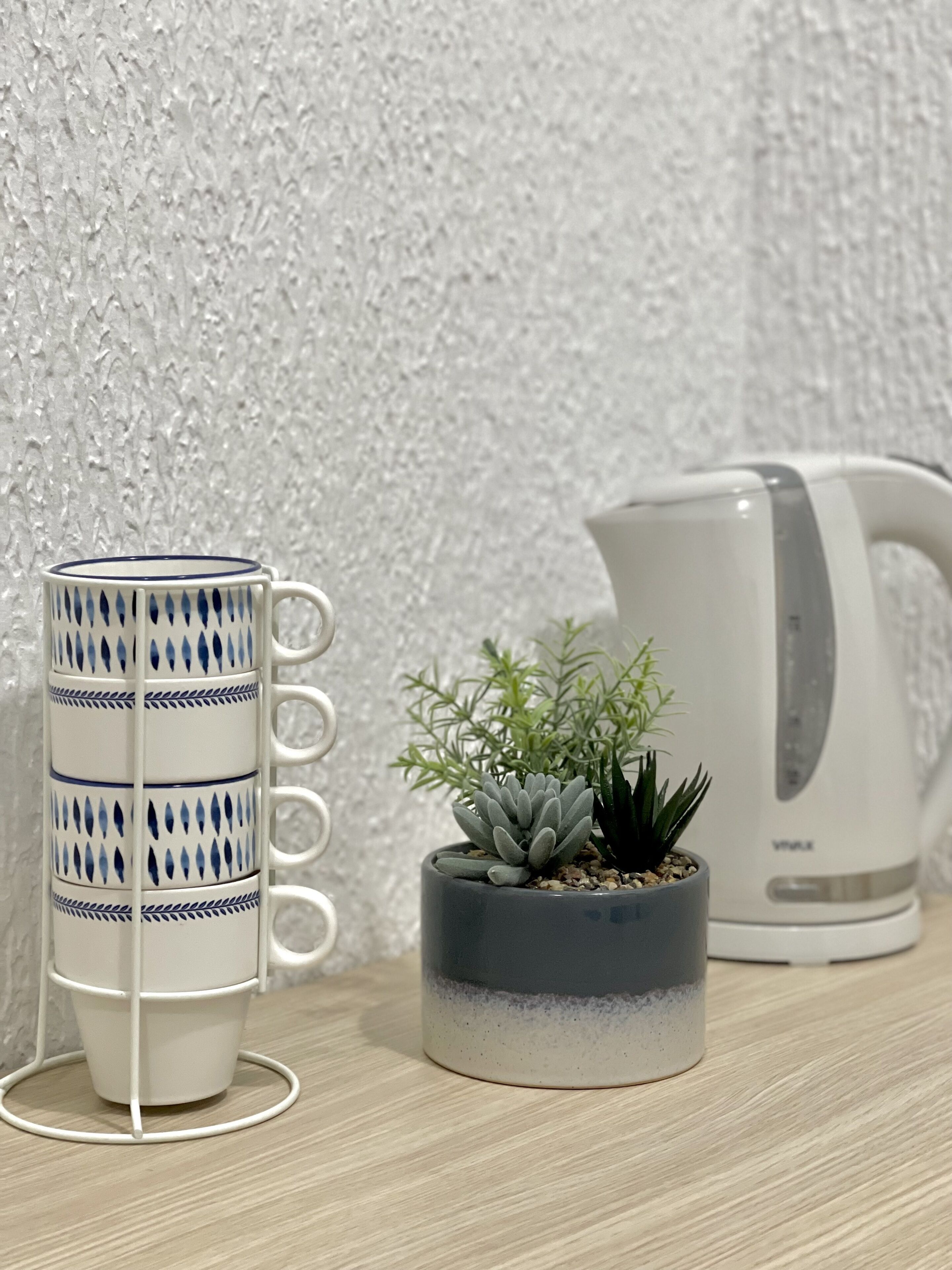 Una pila de tazas blancas con diseños azules, un suculento gris en maceta, y una tetera blanca sobre una superficie de madera frente a una pared con textura.