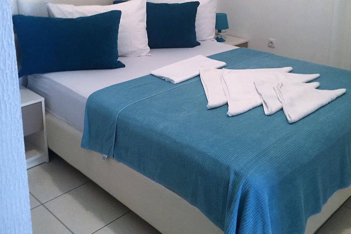 Uma cama bem arrumada com colcha azul e travesseiros brancos, mesa de cabeceira com abajur e cabeceira branca.