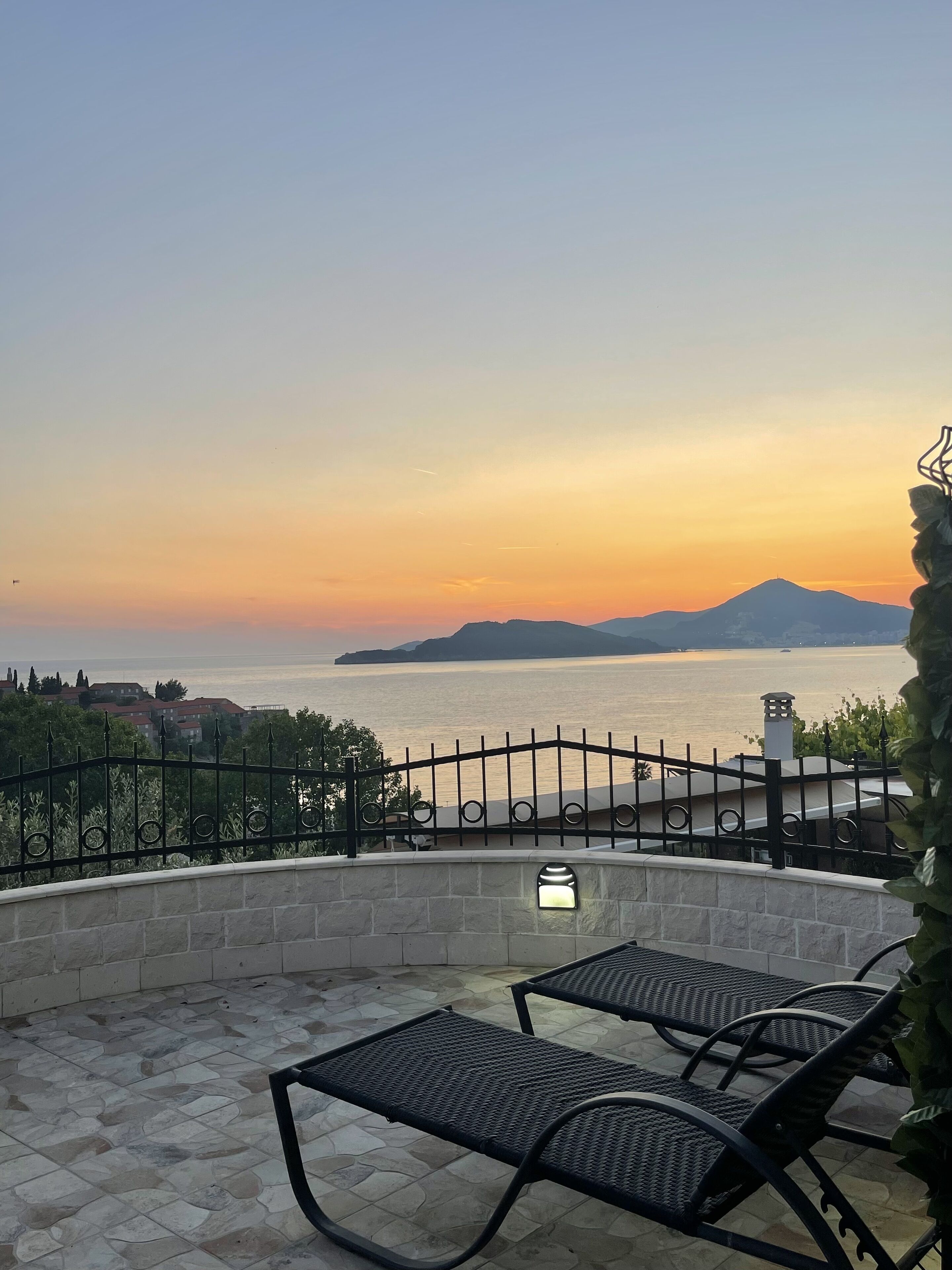 Terraço com duas cadeiras de metal preto, uma parede de pedra e vista para o mar ao pôr do sol.