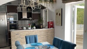 A modern kitchen with a central island, a dining area with blue upholstered chairs and a glass table, and a view through a sliding glass door. - Monte Perla Residence (Sveti Stefan)
