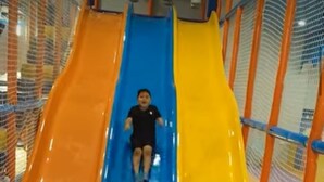 Un niño o niña resbala por un tobogán de muchos colores hacia un pozo de pelotas lleno de pelotas naranja, azul y blanca.
