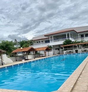 Une piscine avec une terrasse carrelée, un espace de détente couvert et un immeuble à plusieurs étages avec des balcons en arrière-plan.