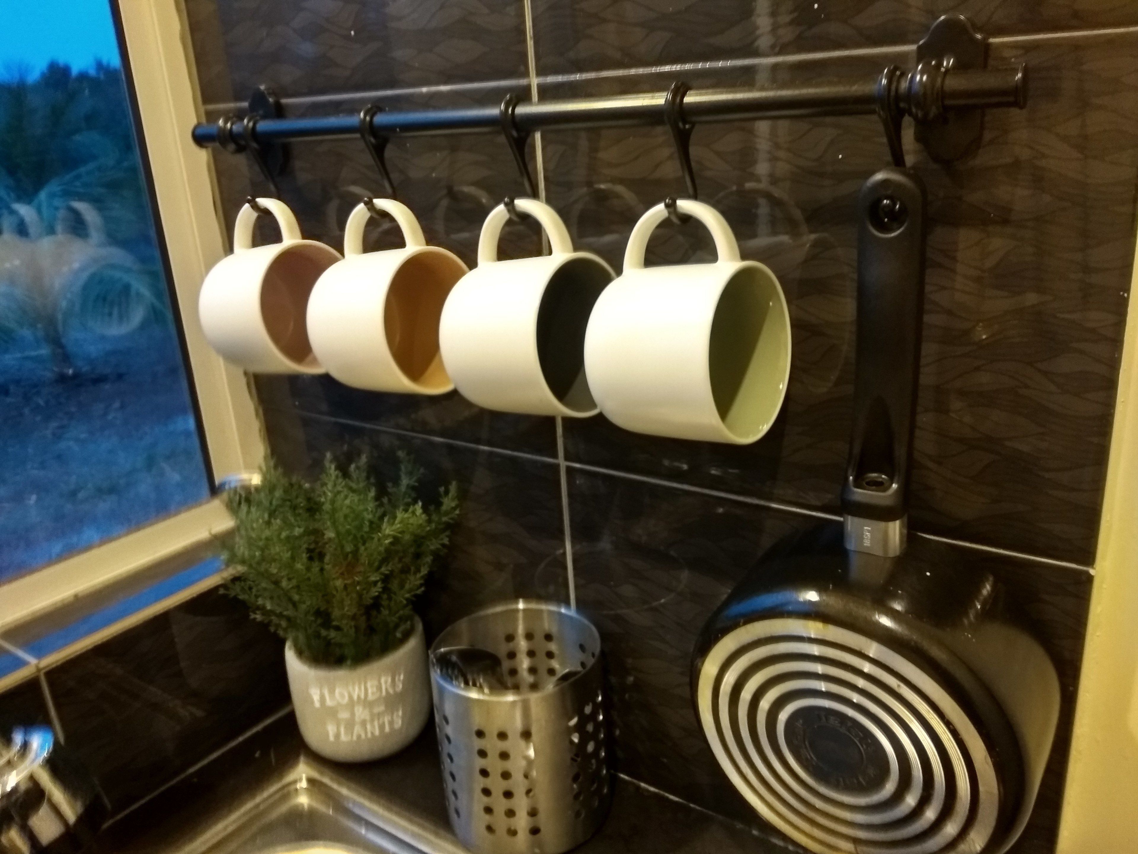 Una cuina amb taulell negre, una barra penjant amb tasses blanques, una planta en test i un colador.