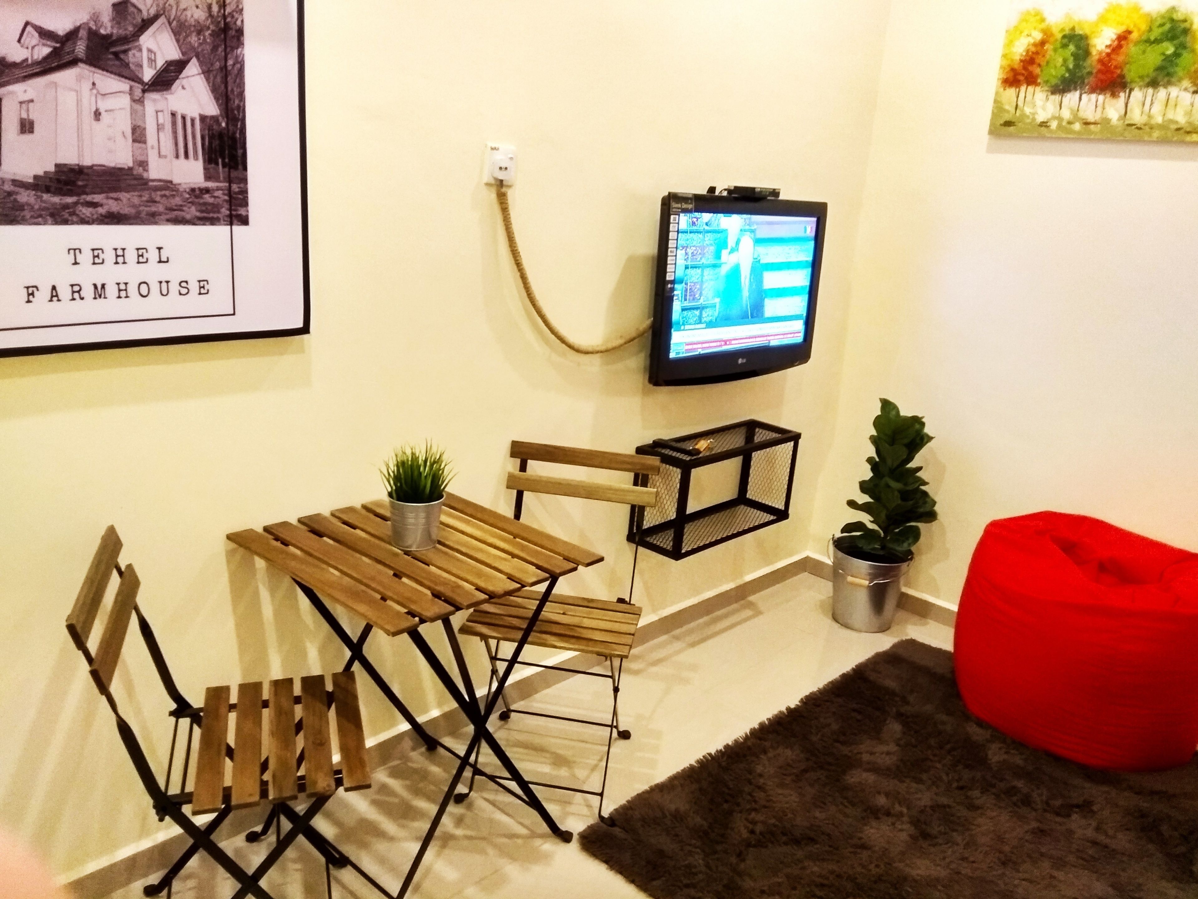 A room with a wooden table and chairs, a television mounted on the wall, a red bean bag, and a framed picture of a farmhouse.