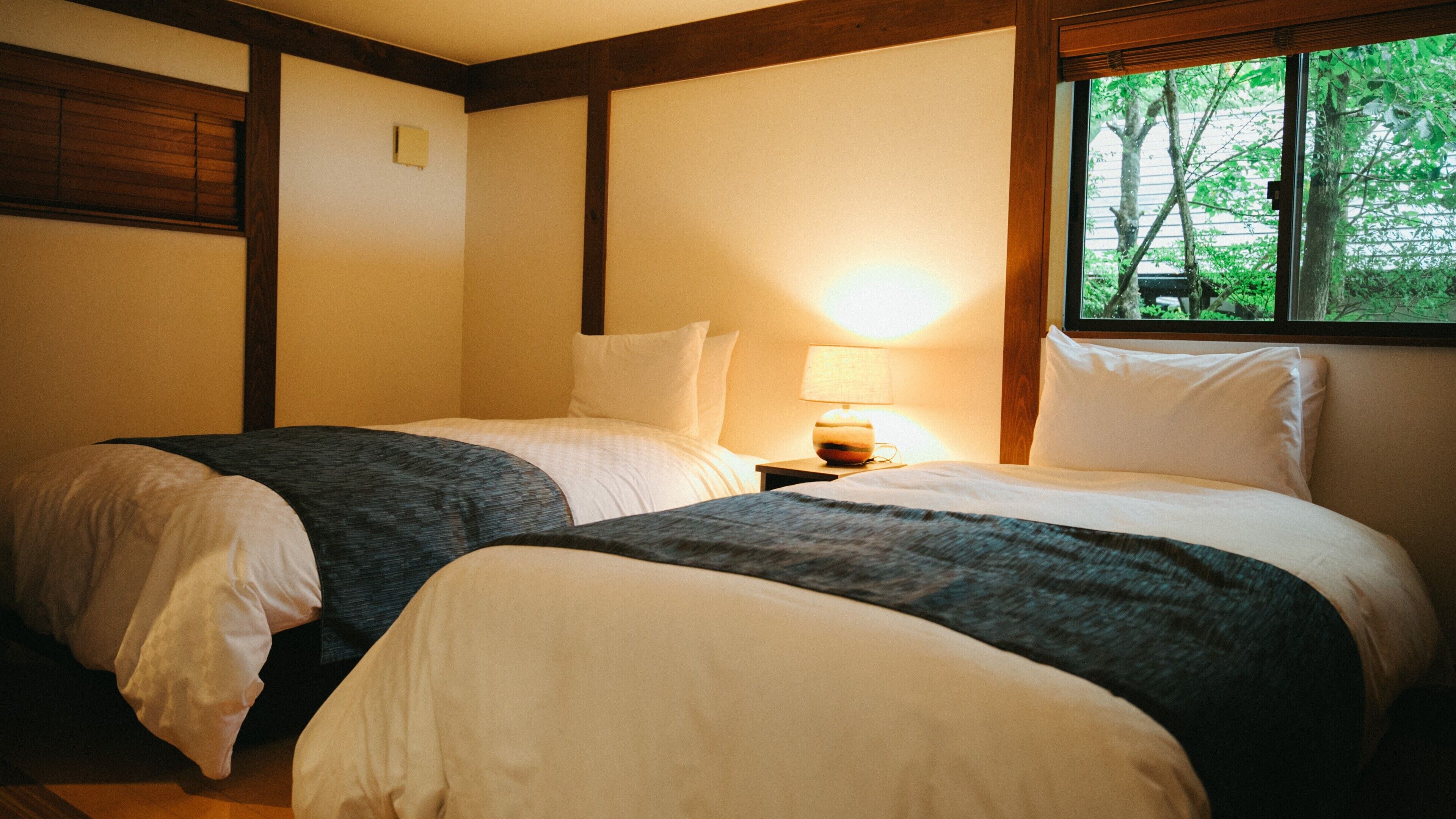 A hotel room with two beds, a nightstand with a lamp, and a window with a view of trees.