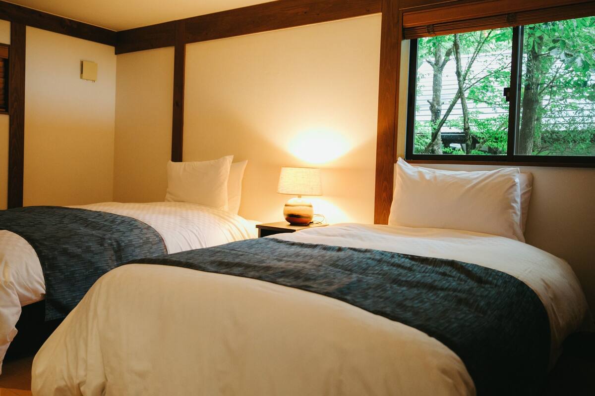 Une chambre d’hôtel avec deux lits, une table de chevet avec une lampe et une fenêtre donnant sur des arbres.