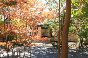 A courtyard with trees displaying autumn foliage, a stone pathway, and a building with a tiled roof. - Auberge - A Ma Façon - (Kokonoe)