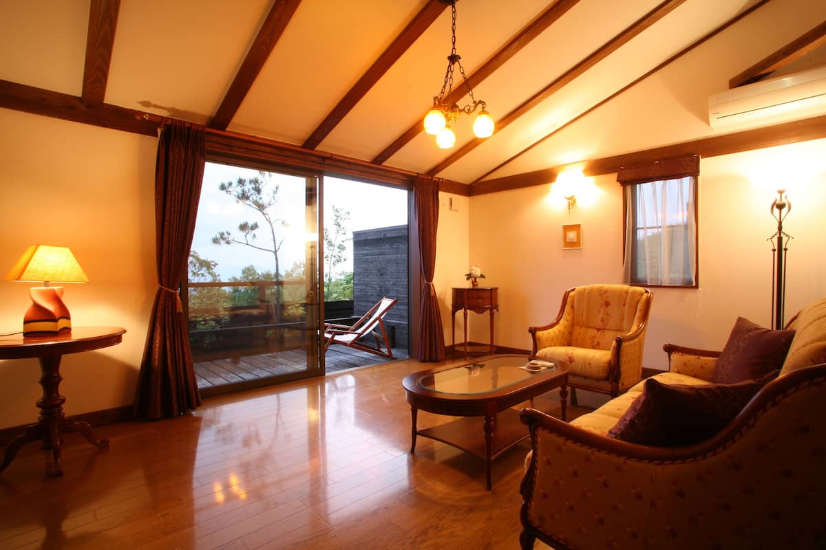Un salon chaleureux avec un plancher en bois, une table basse en verre, un fauteuil à motifs floraux et un canapé. Une porte-fenêtre coulissante donne accès à un espace extérieur.