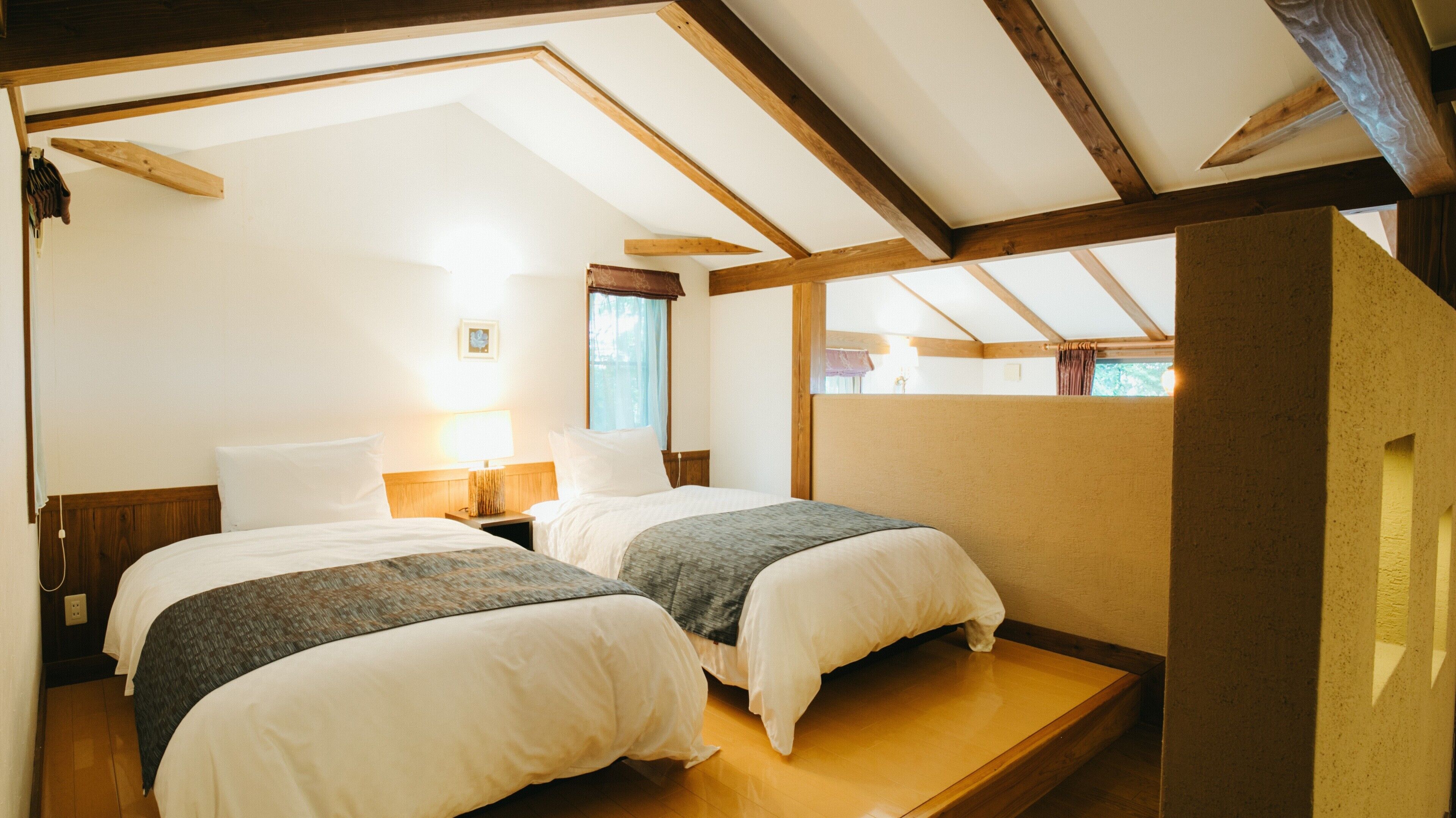 A room with two beds, wooden beams, a window, and a nightstand with a lamp.