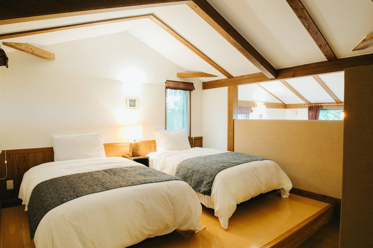 Une chambre avec deux lits, des poutres apparentes, une fenêtre et une table de chevet avec une lampe.