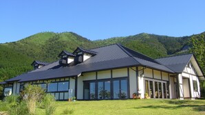 Una casa moderna de dos pisos con ventanas grandes y techo inclinado, rodeada de abundante vegetación y colinas.