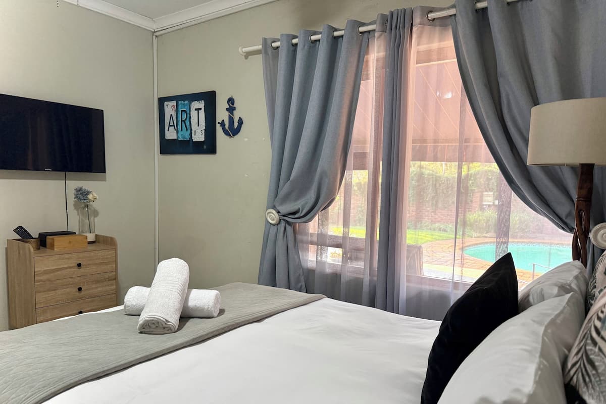 A neatly made bed with white linens, a wooden nightstand with a vase and remote control, a wall-mounted TV, and a view of a swimming pool through large windows with grey curtains.