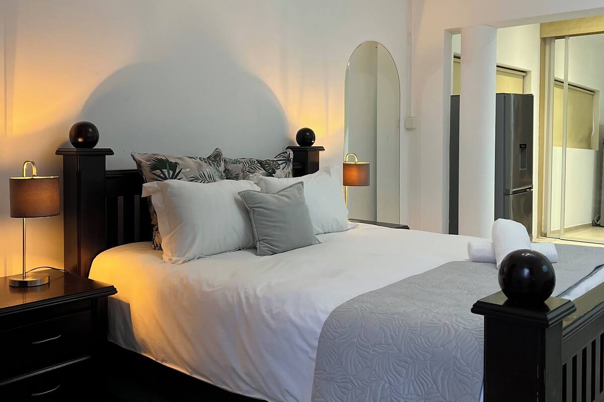 A neatly made bed with a white comforter and pillows, a bedside table with a lamp, a wall-mounted air conditioner, and a mirror above the bed.