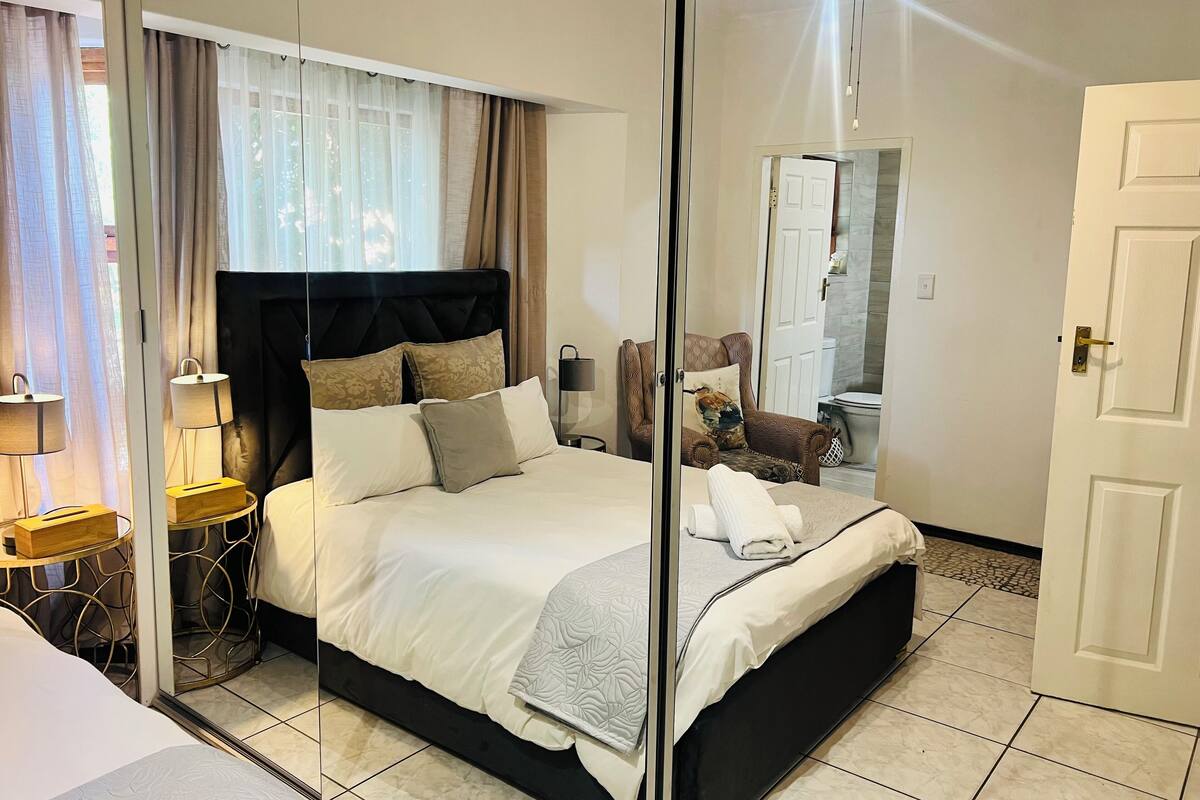 A hotel room with a large bed, a bedside table with a lamp, a chair with a cushion, and a mirror reflecting the room.