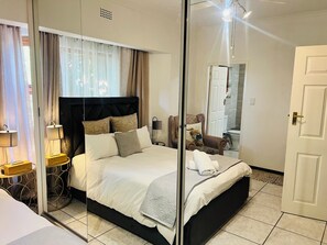 A hotel room with a large bed, a bedside table with a lamp, a chair with a cushion, and a mirror reflecting the room. - Map's View Guesthouse (Johannesburg South)