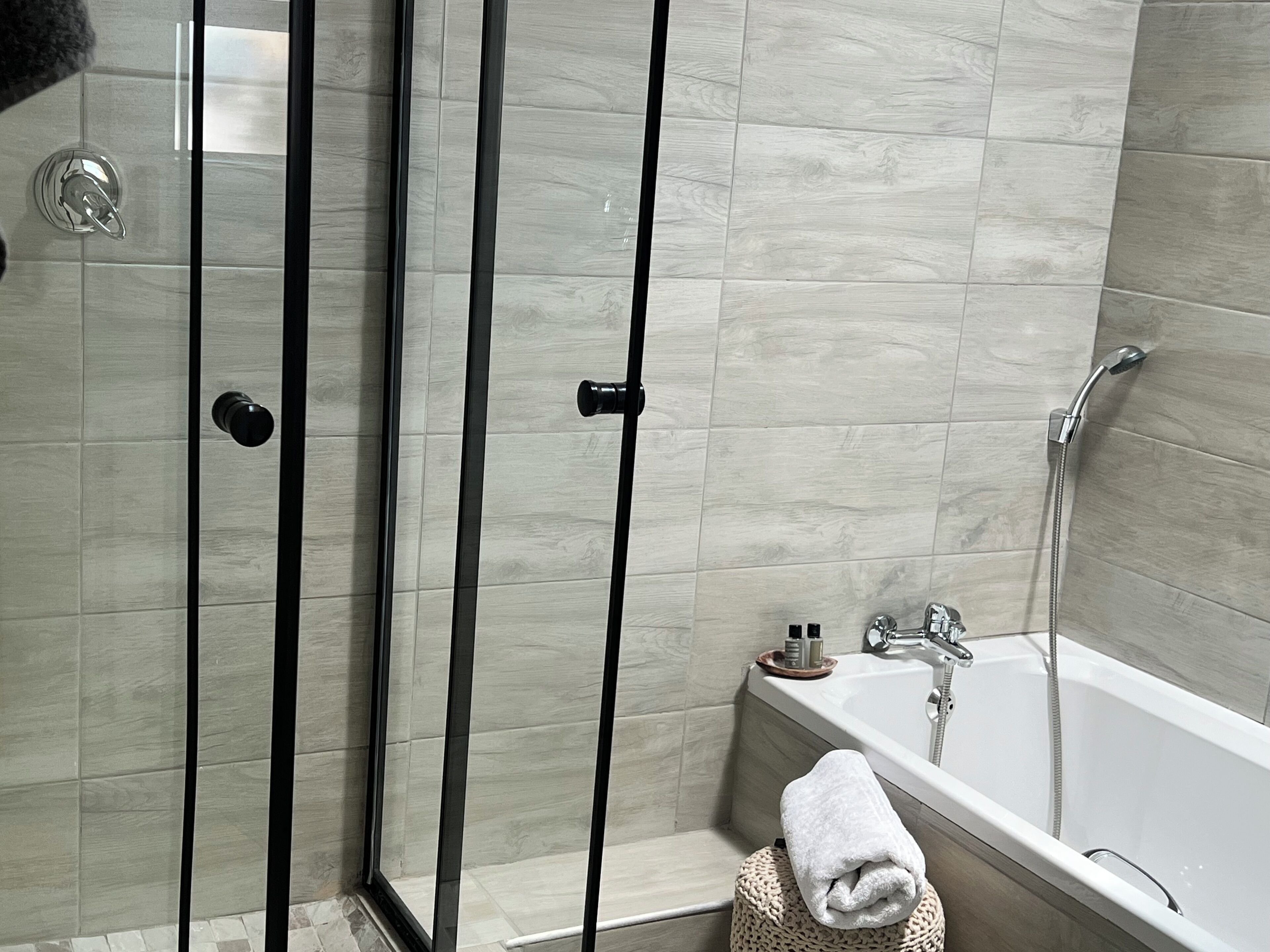 A modern bathroom with a glass shower enclosure, a bathtub, and a small bedside table with toiletries.
