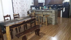 Un coin repas avec une table et des bancs en bois, une cuisine avec des meubles sombres et un réfrigérateur, et un rideau à motifs.